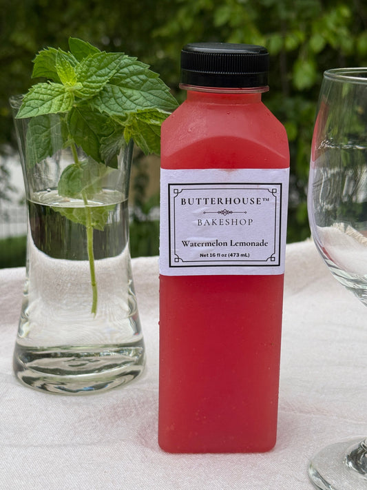 Bottle of Butterhouse Bakery watermelon lemonade with a glass of water and a mint leaf on a white cloth.